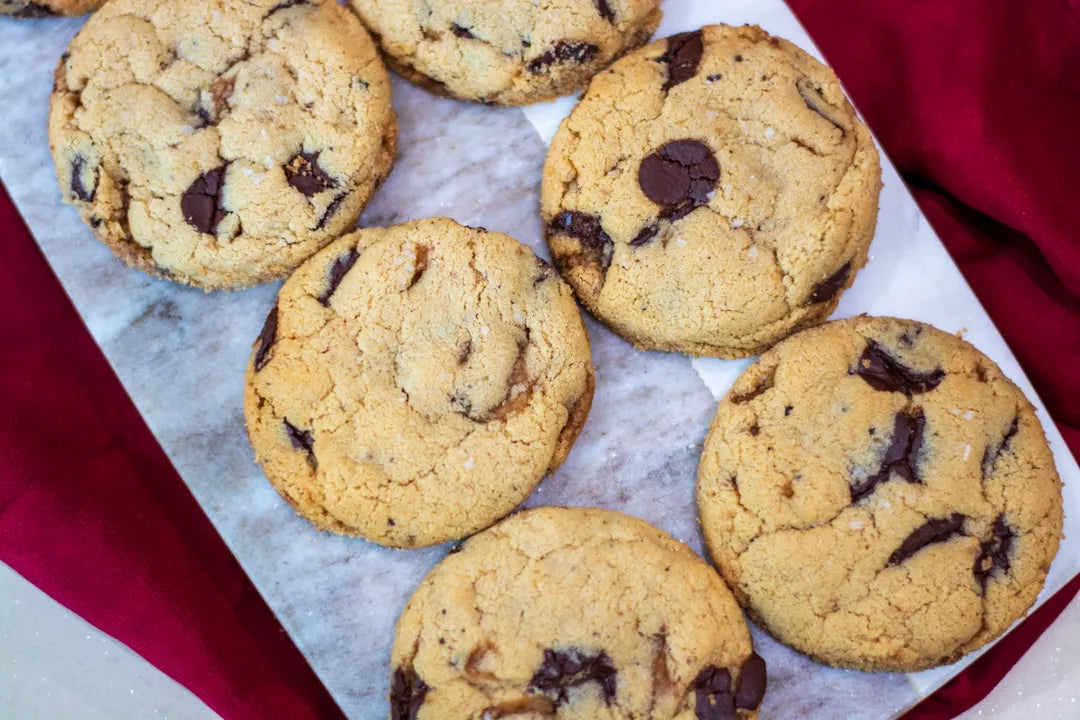 Six golden-brown gluten-free chocolate chip cookies arranged on a white marble serving board with a red cloth underneath.