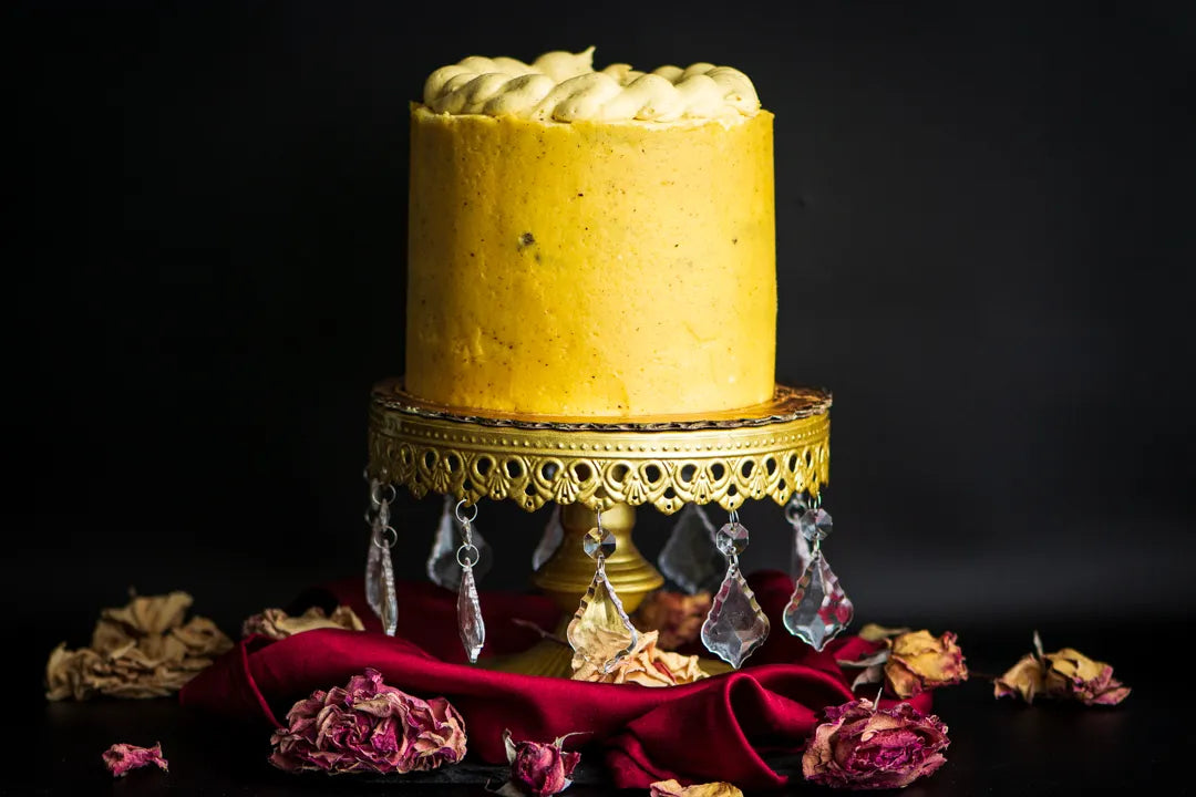 A decorated gluten-free caramel cake with a golden yellow-hued frosting, placed on a cake stand with a red velvet cloth and adorned with crystal drops and caramel pieces on the sides.