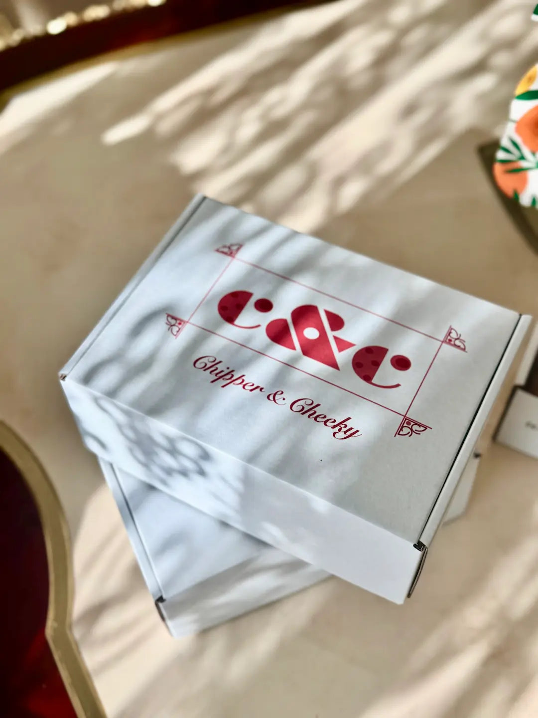 A white product box with red printed branding reading 'c&c' and 'Chipper & Cheeky' gluten-free bakery sits on a light beige surface with soft shadow patterns across it.
