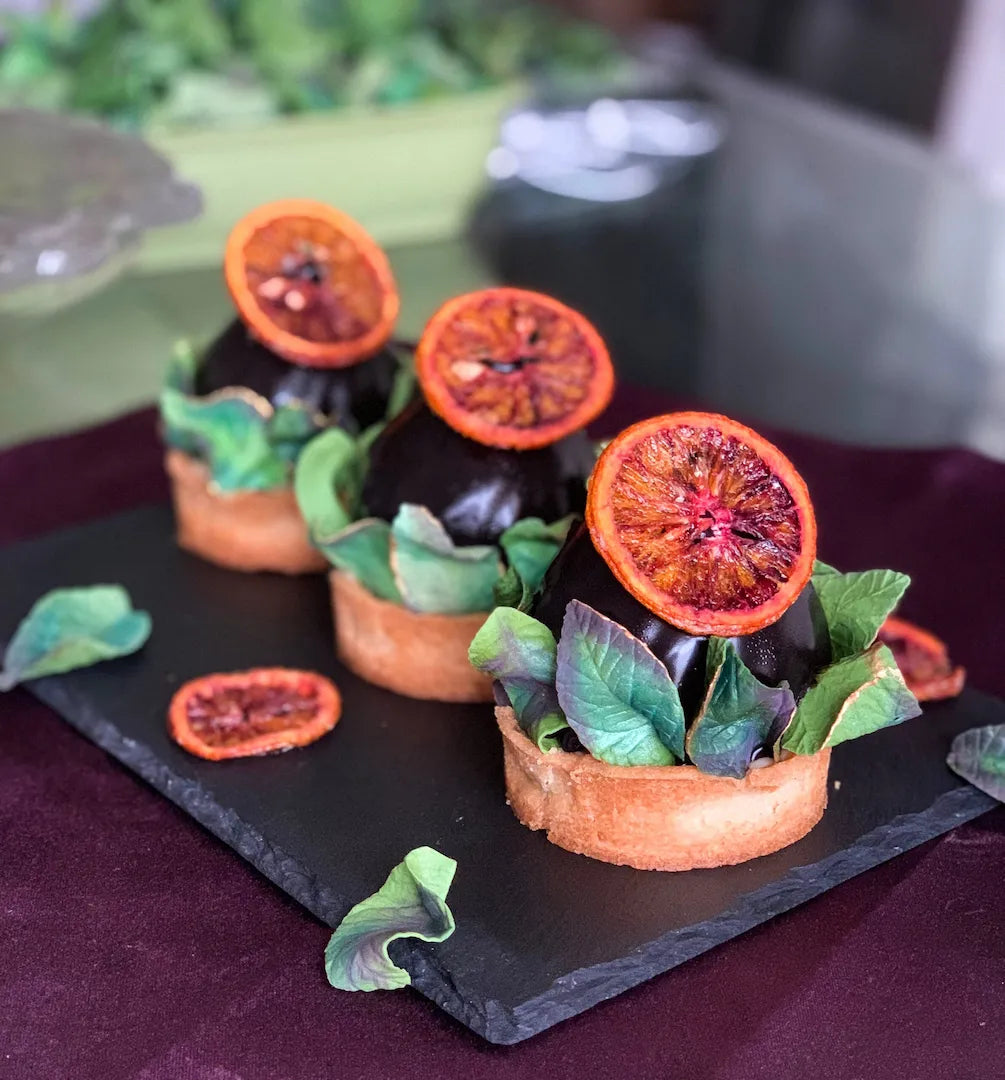 Three small gluten-free chocolate-blood orange tarts for a wedding in Valencia California, topped with sliced blood oranges and green leaves, arranged on a black serving platter over a dark purple cloth.