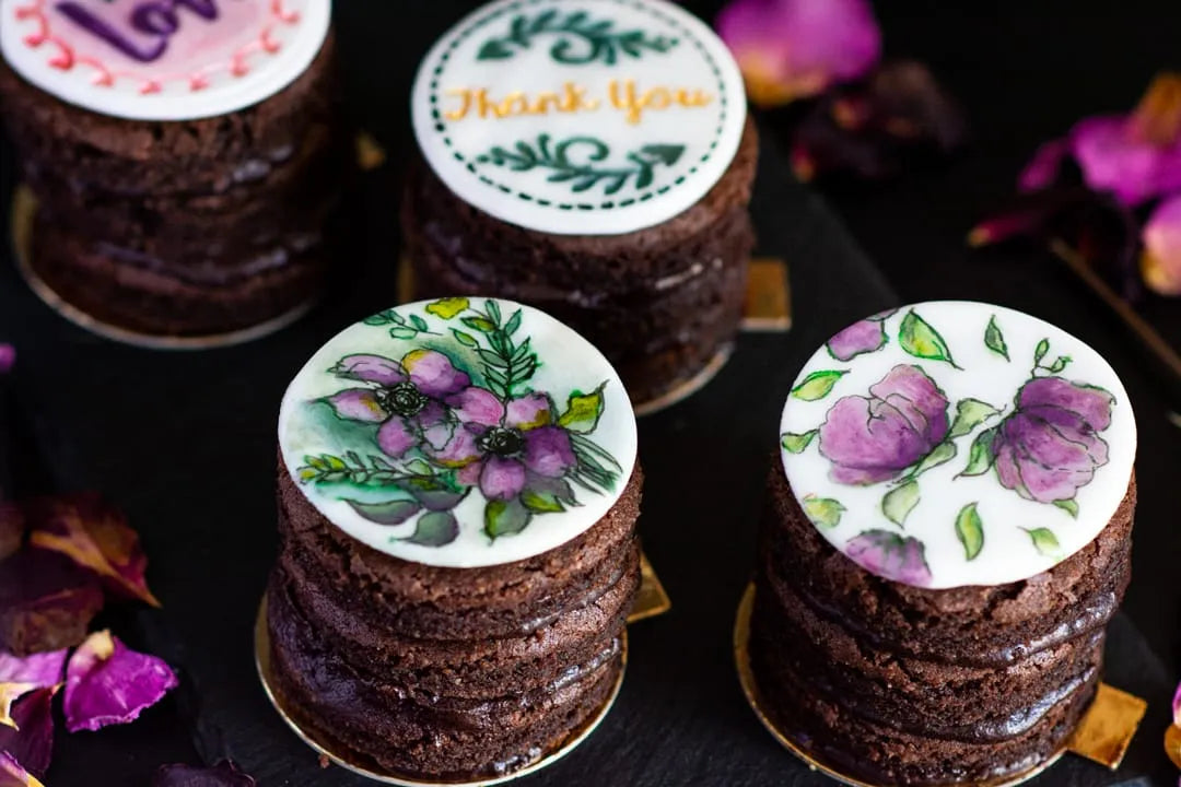 Four small gluten-free chocolate cake stacks for a Santa Monica bridal shower, decorated with round white topper lids featuring floral designs and one with a 'Thank You' message, arranged on a dark surface with dried rose petals.