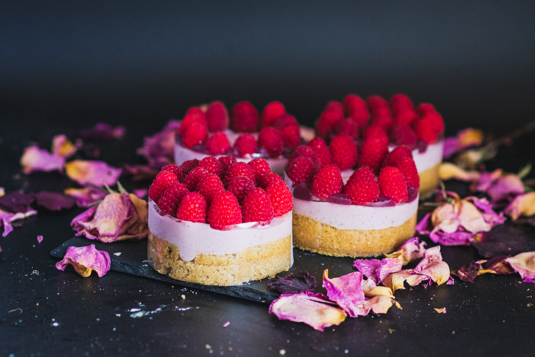 A minimalist luxury dessert spread displaying four individual Chipper & Cheeky gluten-free raspberry mousse mini cakes topped with fresh local raspberries, optimized for high-end dessert tables and corporate gifting.