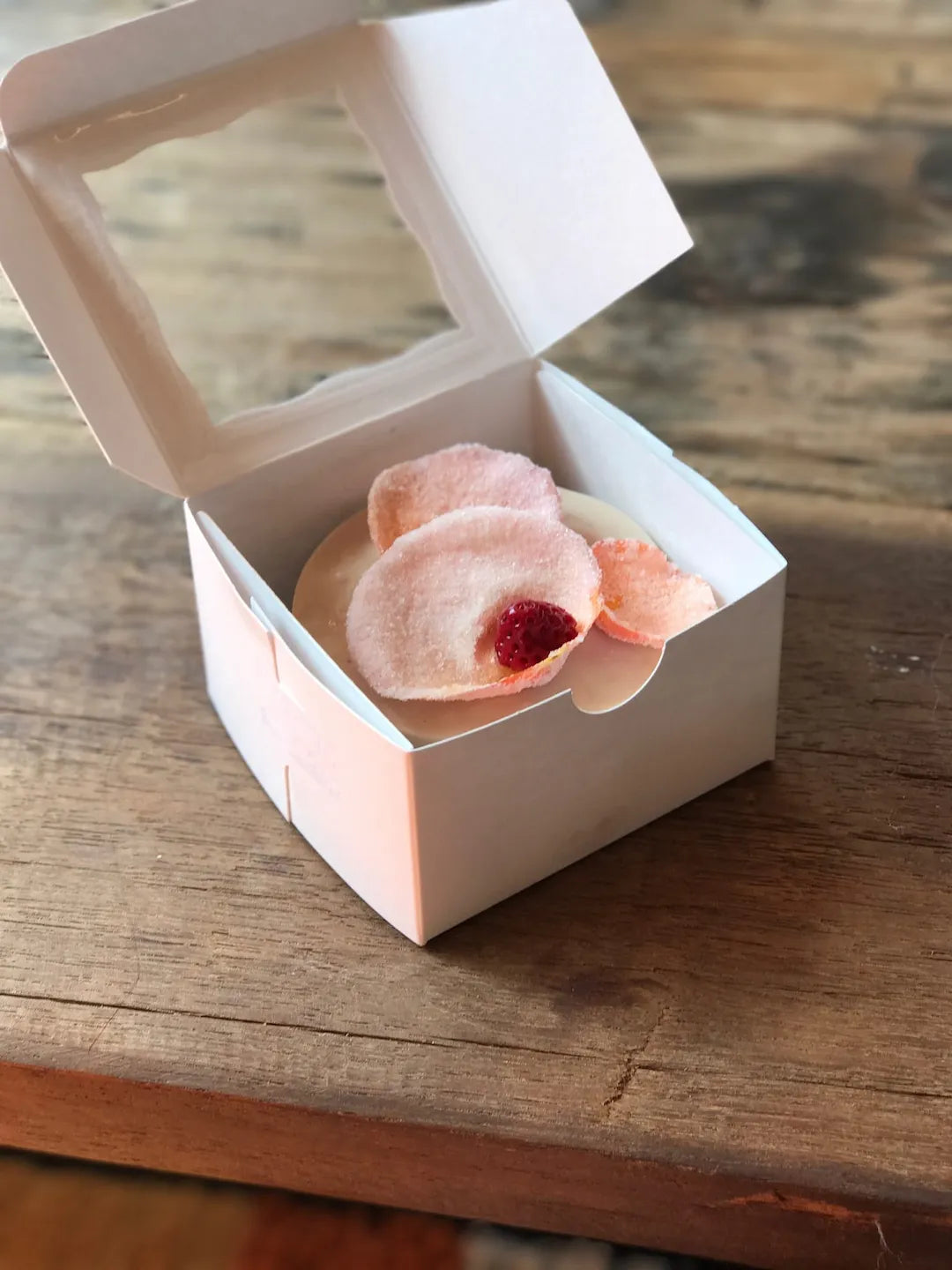 A small white paper food box with a clear window lid, open to reveal a gluten-free rose cake topped with pale pink petals and a dark red berry, set on a rustic wooden table.