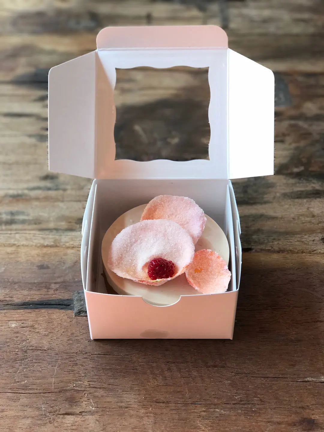 A pink paper bakery box with a clear window in the lid, containing a small gluten-free mini rose cake topped with pink sugar glaze and a raspberry, set on a rustic wooden table.