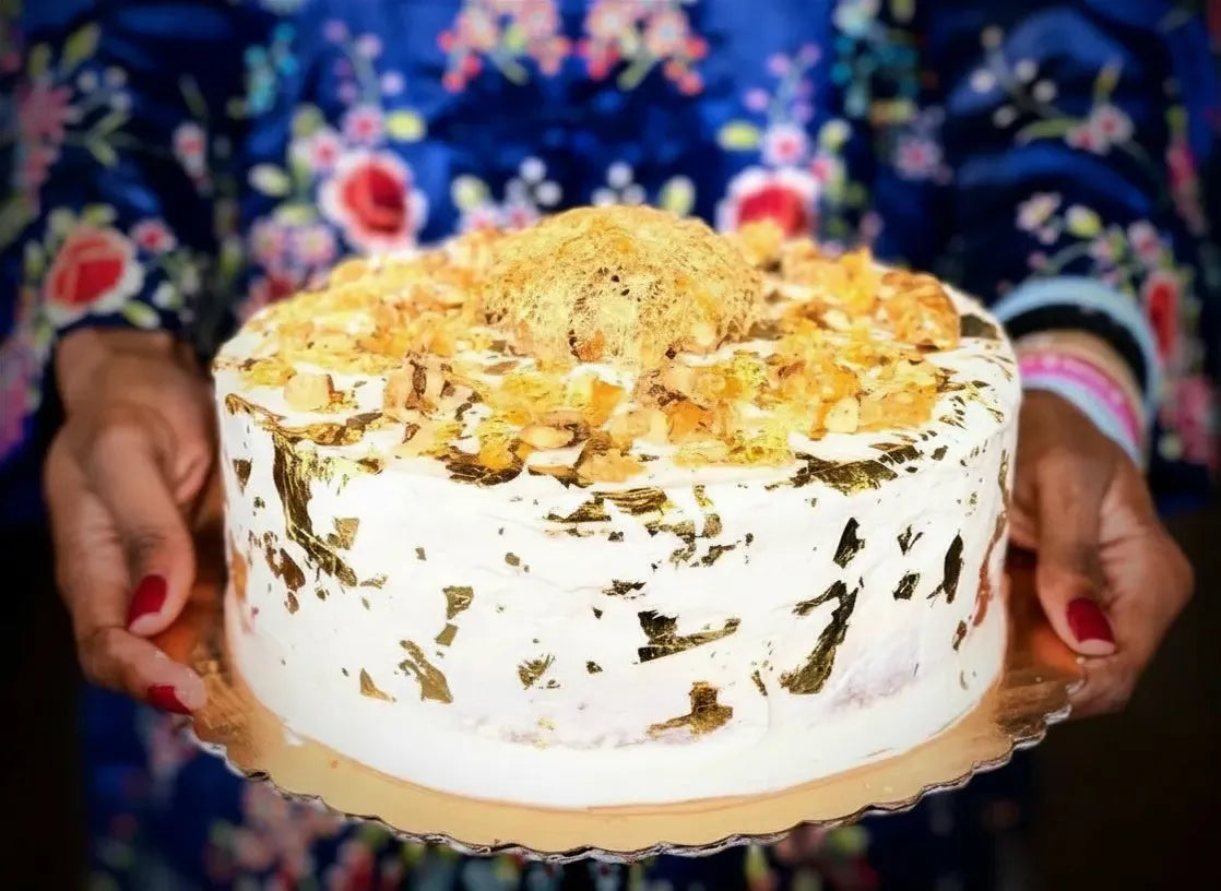 A round white frosted gluten-free sweet potato cake topped with golden flakes and a golden spun sugar cage, held by hands against a colorful floral blue background.