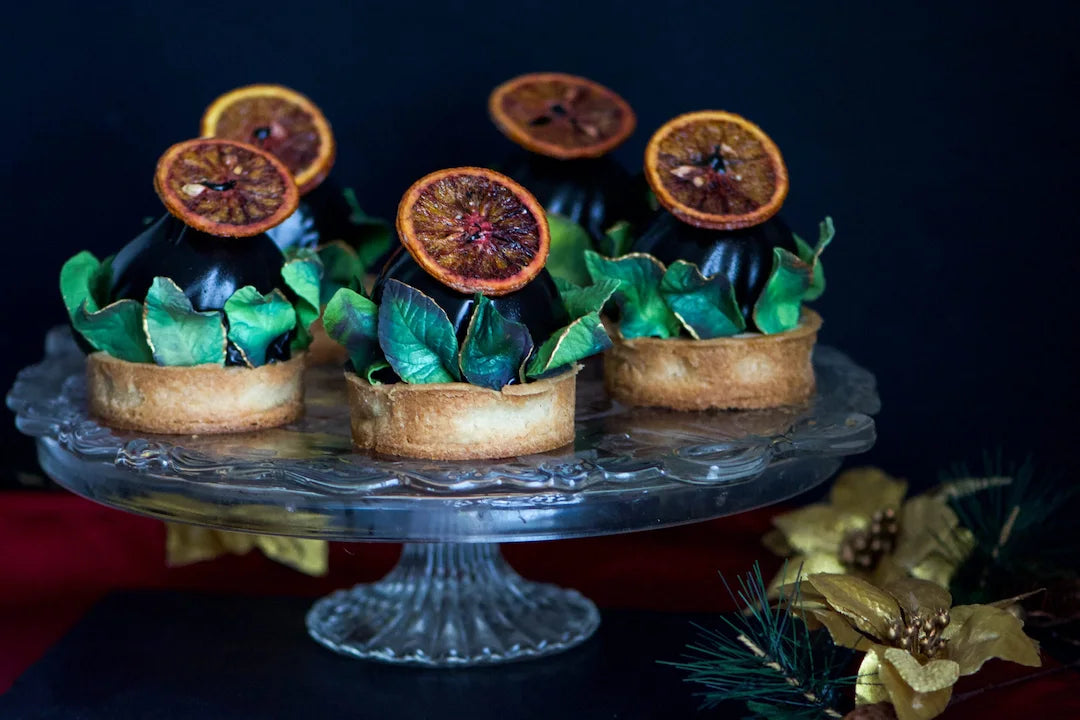 Three small gluten-free tartlets for a Los Angeles private event, filled with dark chocolate and topped with dried citrus slices and green leaves, displayed on a clear glass pedestal plate against a dark background.