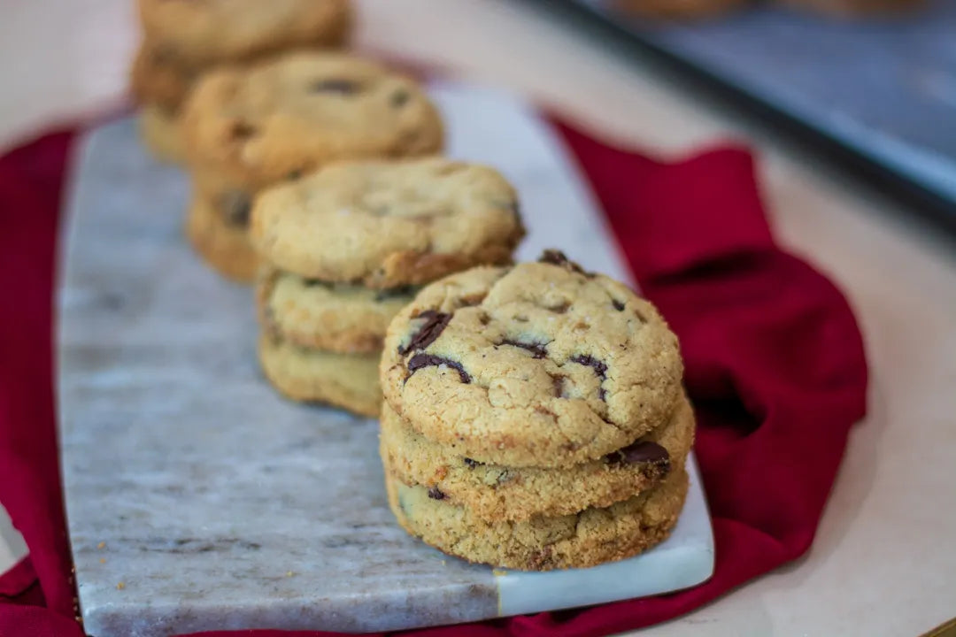Three stacked gluten-free chocolate chip cookies on a white marble serving board, with additional cookies blurred in the background and a red cloth underneath.