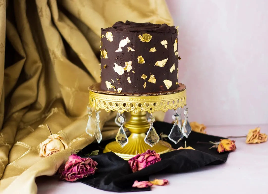 A gluten-free chocolate-covered cake with gold leaf flakes sits on a gold pedestal stand decorated with hanging clear crystals, styled with gold fabric, black cloth, and dried roses.