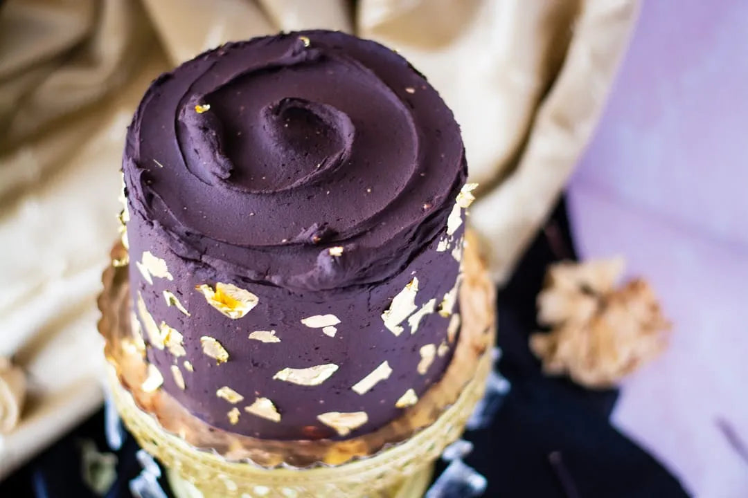 A close-up of a frosted gluten-free cake with dark chocolate icing, swirled on top and decorated with scattered gold flakes, set against a soft beige and lavender fabric background.