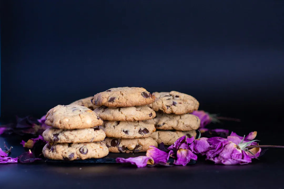 A stack of gluten-free hazelnut chocolate chip cookies on a dark surface with purple flower petals around them, set against a dark blue background.