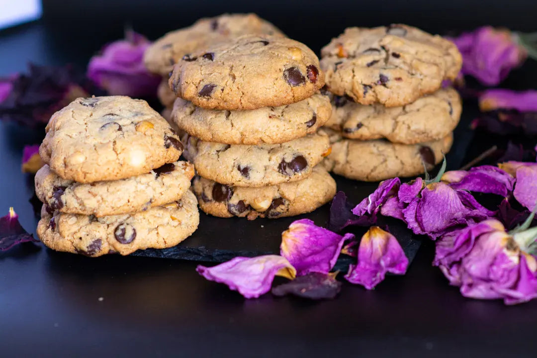 Three stacks of gluten-free hazelnut chocolate chip cookies on a black slate plate, surrounded by purple dried flower petals on a dark background.