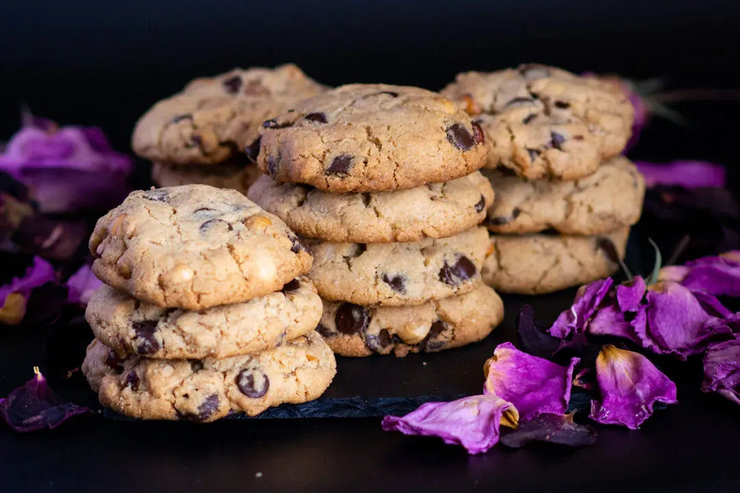 Stacks of golden-brown gluten-free chocolate chip cookies on a dark surface, surrounded by scattered purple flower petals.