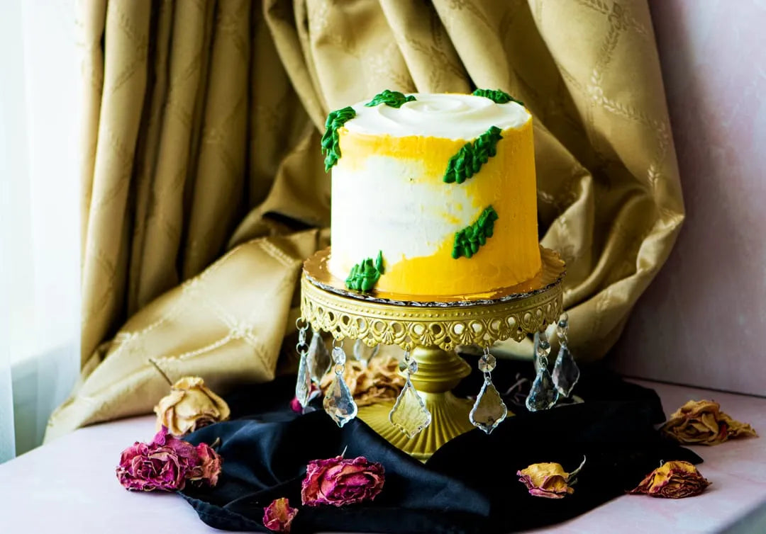 A frosted gluten-free lemon cake with yellow and white icing, decorated with green leaf-like piped details, displayed on an ornate gold cake stand with hanging crystal drops. The stand sits on a black cloth with scattered dried roses, set against draped gold fabric.