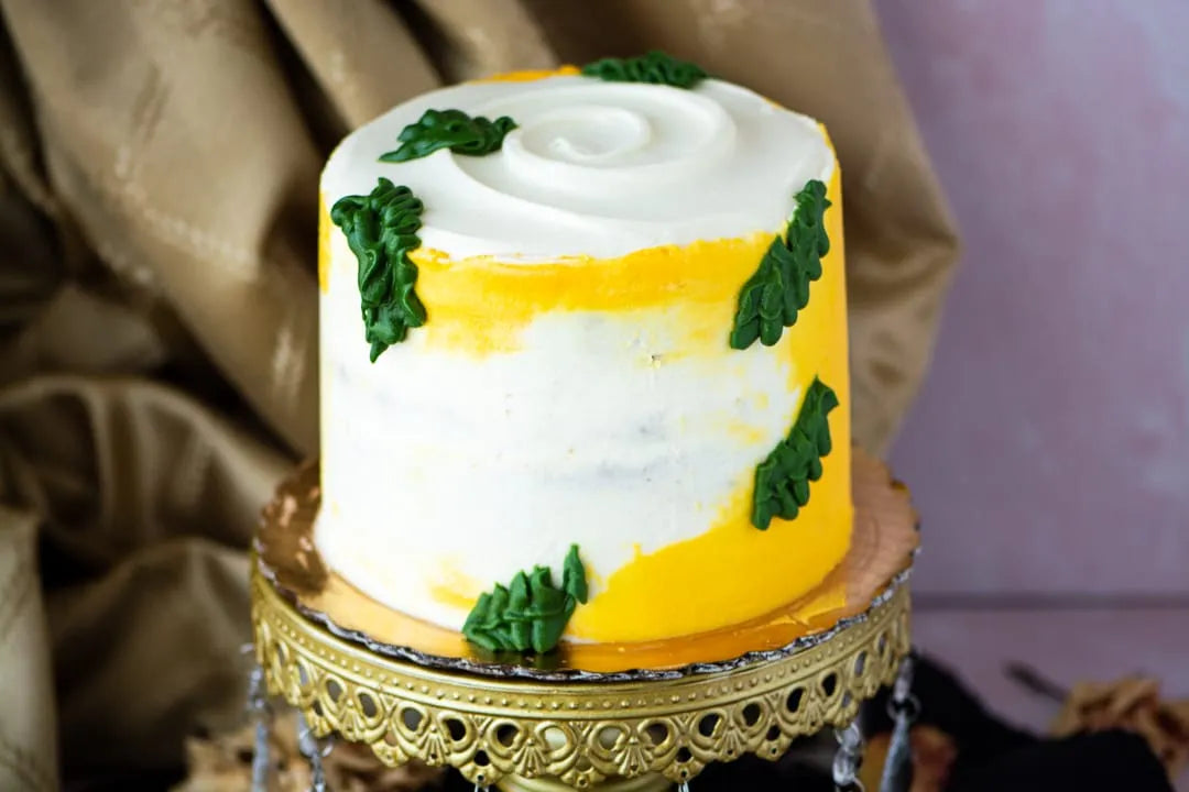 A round frosted gluten-free lemon cake with a white top, yellow side bands, and green leaf decorations, displayed on an ornate gold pedestal against a draped fabric background.