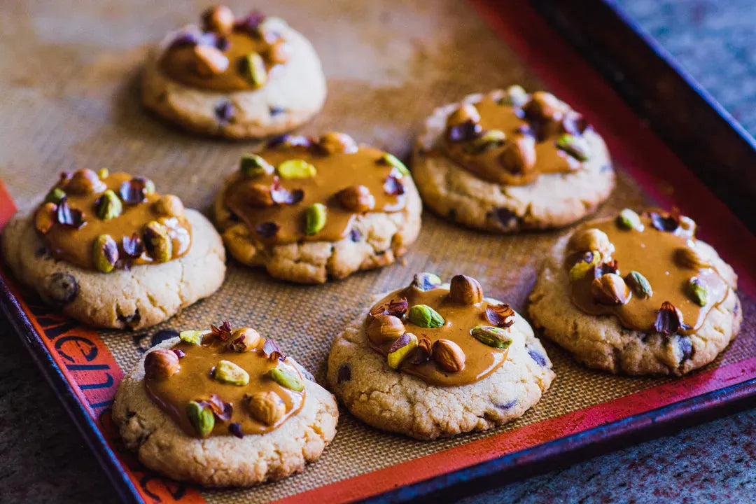 Seven gluten-free chocolate chip cookies for a Los Angeles shipped order, topped with freshly made praline, nuts, and pistachios, arranged on a baking sheet lined with a silicone mat.