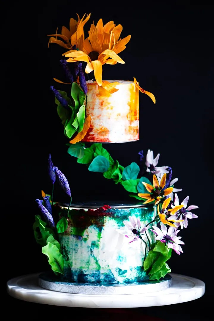 A decorative two-tier wedding gluten-free cake displayed on a white round stand against a black background. The cake features a lower tier with blue-green marbled frosting and edible sugar green leaf decorations, a middle tier with orange marbling on white, and a top tier shaped like a candle in white with orange marbling.