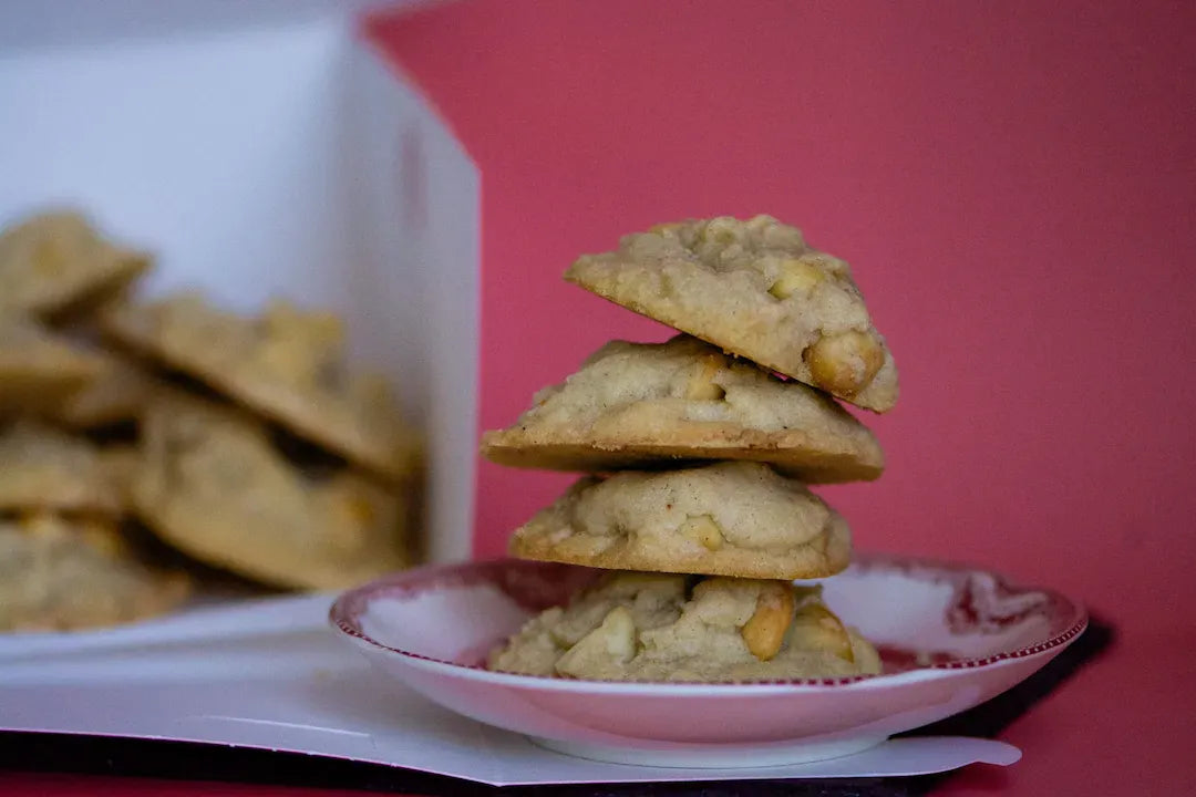 A stack of four golden vegan, gluten-free Macadamia Nut cookies for a private event in Los Angeles, made with nuts and white chocolate, served on a small pink plate with a red rim, with more cookies blurred in a white box in the background against a pink backdrop.