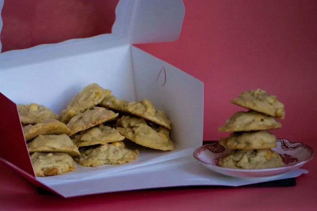 A white paperboard box filled with golden-brown vegan, gluten-free white chocolate macadamia nut cookies, with a small stack of cookies on a pink-and-white plate beside it, set against a solid pink background, for a corporate event in Valencia California.