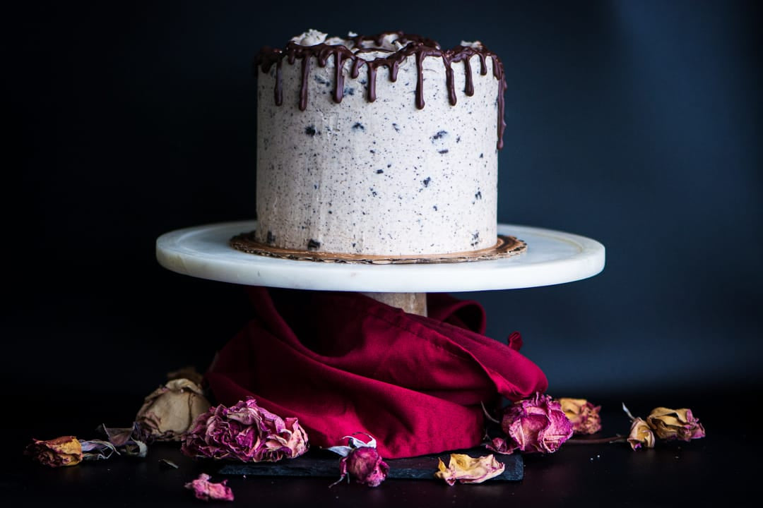 A frosted gluten-free cake with dark chocolate drizzle sits on a white round cake stand, with a deep red fabric drape and scattered dried pink roses below, against a dark background.