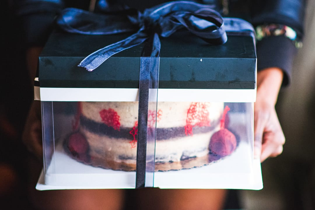 A half-snickered cake with red berry garnish inside a clear plastic box with a black lid and ribbon, held by hands against a dark background.