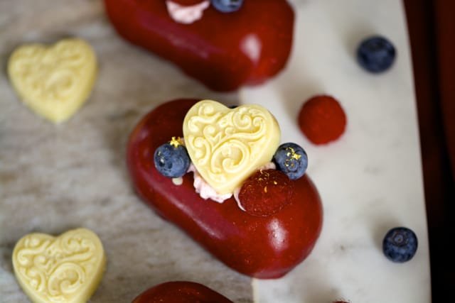 Heart-shaped white chocolates topping a gluten-free, red-colored entremet, arranged on a marble surface with additional heart-shaped chocolates and berries.
