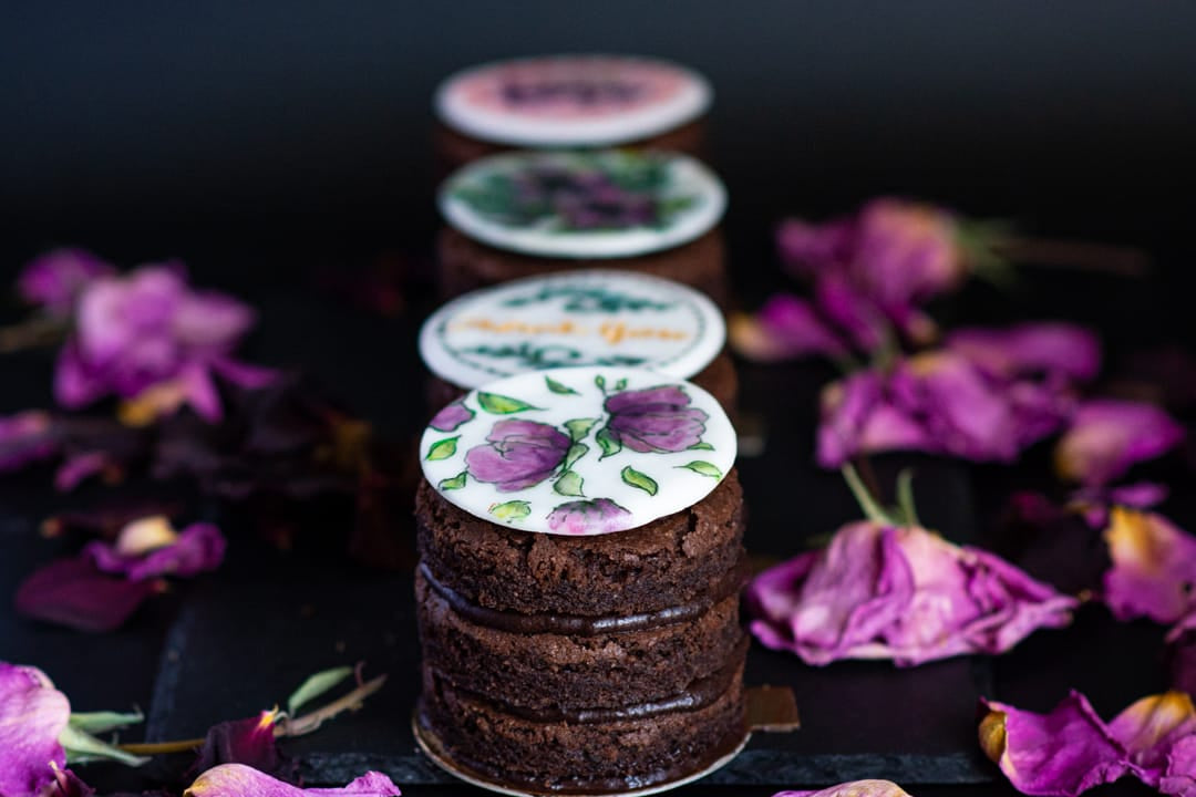 Four small gluten-free chocolate cake layers for a Los Angeles birthday, stacked vertically and decorated with round white toppers featuring purple rose illustrations and cursive text, arranged on a dark surface with scattered dried purple flowers.