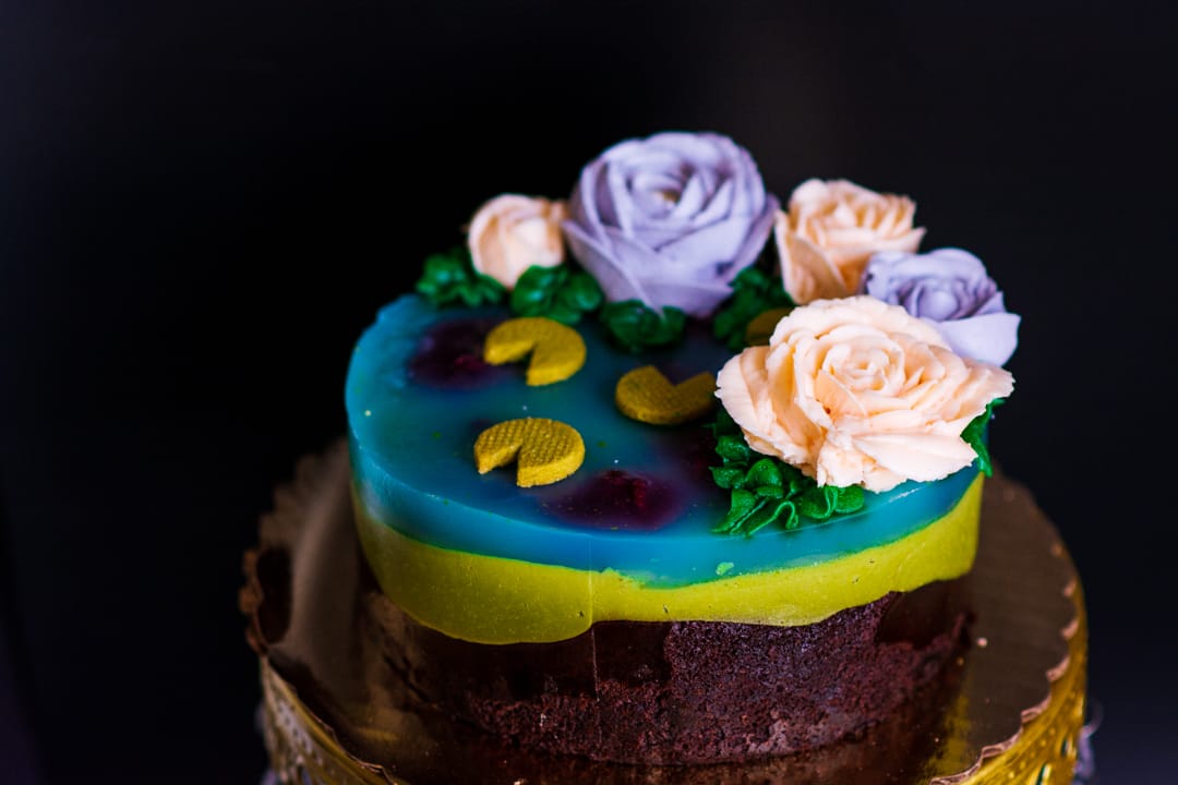 A decorated gluten-free chocolate cake on a gold-rimmed stand, featuring a blue and yellow top layer with piped purple and peach roses, green leaves, and gold crescent moon decorations against a dark background.