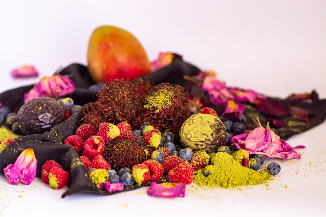 A styled botanical and fruit arrangement on a white background, featuring raspberries, blueberries, a mango, dried pink flower petals, dark fabric, and bright yellow-green powder and seeds.