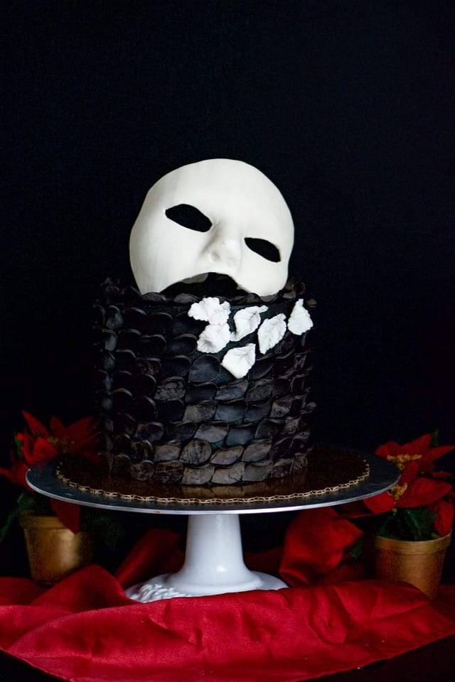 A black gluten-free chocolate cake with a textured, scale-like frosting pattern, topped with a white fondant mask and decorated with white flower shapes, displayed on a white cake stand against a black background with red fabric and poinsettias.