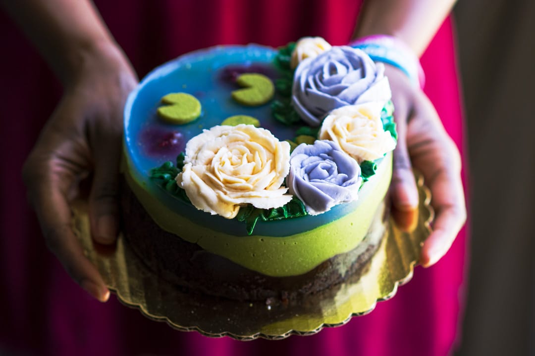 A round decorated cake held in two hands. The cake has a blue top with lily pad designs, cream and lavender roses, green frosting accents, and a dark brown sponge layer visible on the sides.