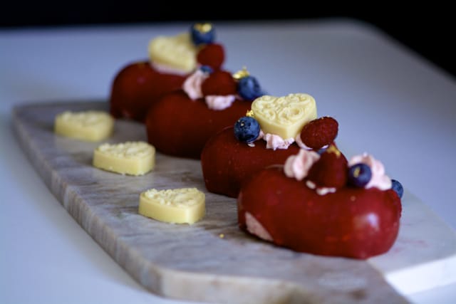 Four red heart-shaped gluten-free pastries topped with cream, raspberries, blueberries, and heart-shaped white chocolate pieces, arranged on a marble serving board.
