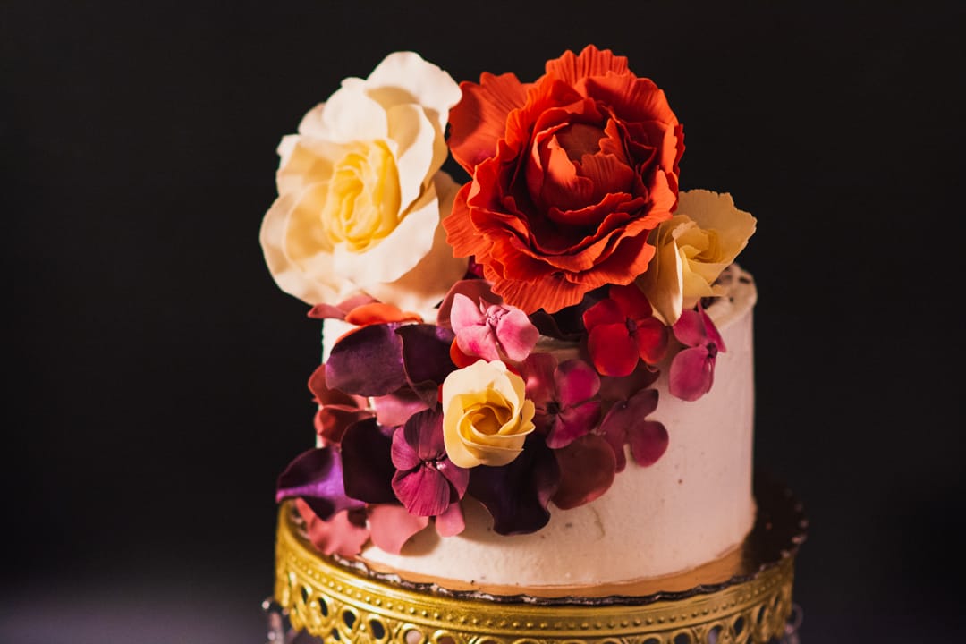 A two-tier gluten-free white frosted cake decorated with large artificial flowers in red, cream, yellow, and purple, displayed on a gold pedestal against a dark background.