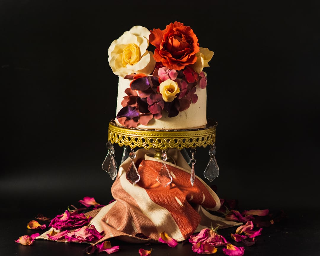 A two-tiered white cake decorated with large artificial flowers in red, yellow, and purple tones, displayed on an ornate gold pedestal with hanging clear crystals against a black background.