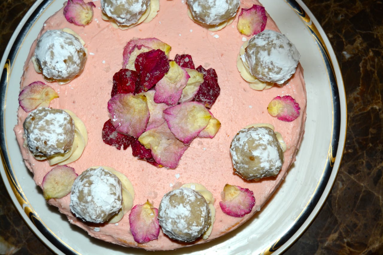 A pink frosted gluten-free cake topped with small powdered white chocolate truffles, crystallized rose petals, and a central cluster of dark red and yellow crystalized flower petals, served on a white plate with a gold rim.