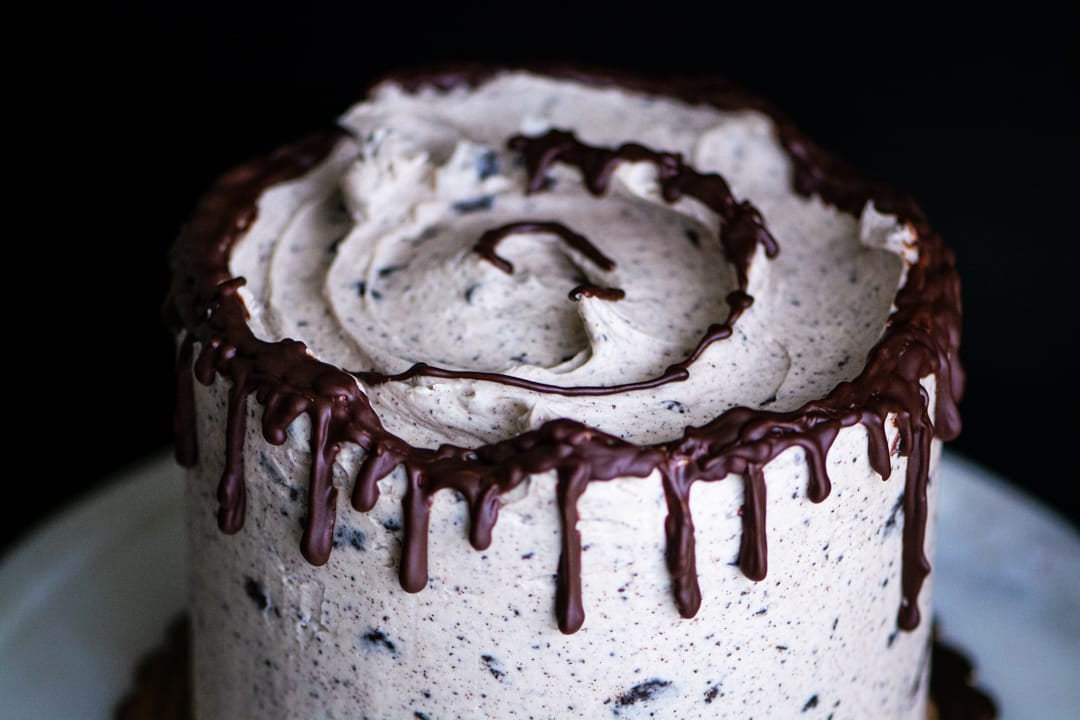 A close-up of a frosted  gluten-free cake with white icing speckled with dark cookie bits and topped with dark chocolate drizzle, set against a black background.