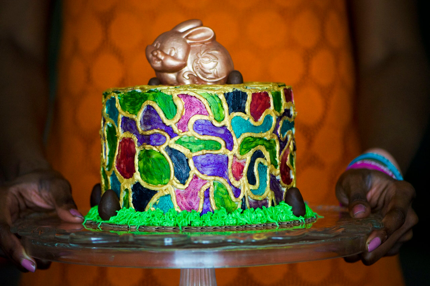 A decorated Easter gluten-free cake with a gold chocolate bunny topper, held on a glass cake stand by hands against a warm orange patterned background.