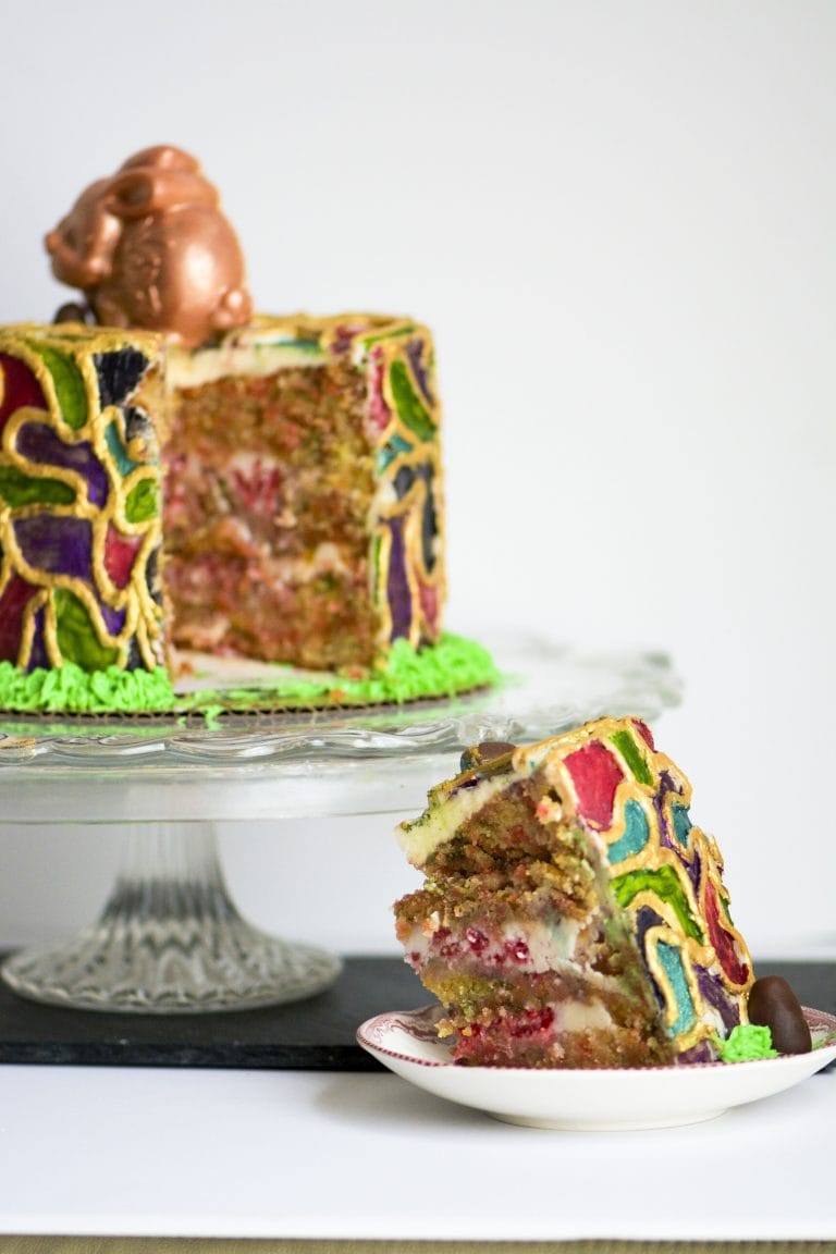 A decorated gluten-free funfetti cake with a slice cut out and placed on a small plate in front, set on a glass cake stand against a plain light background. The cake has a colorful mosaic-style frosting pattern in green, yellow, red, and purple, with a chocolate pig decoration on top.