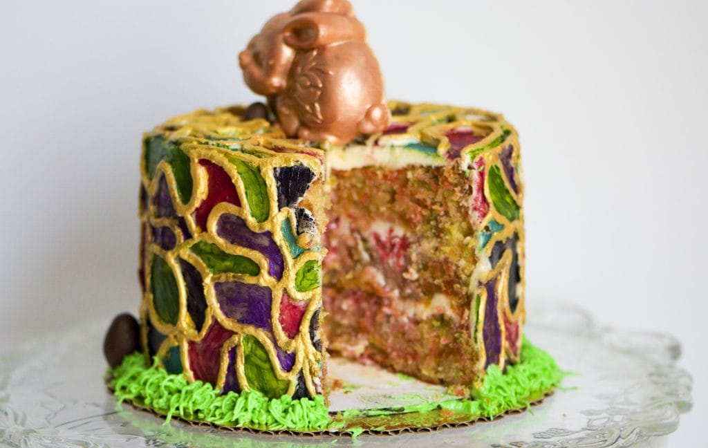 A decorated gluten-free cake with a cut slice showing the interior, topped with a small chocolate bunny. The cake is covered in colorful mosaic-like icing with gold outlines and green, purple, and blue sections, set on a clear glass plate against a plain light background.